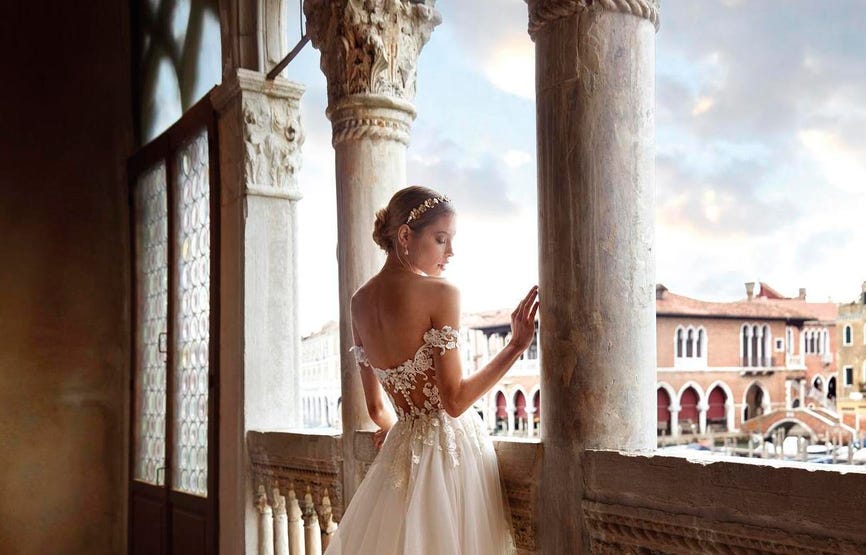 Modella di spalle che si affaccia da un porticato e si guarda la mano mentre indossa un abito da sposa con maniche pendenti.